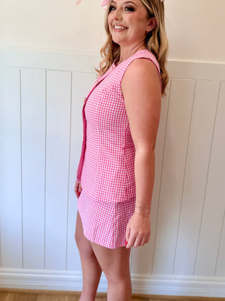 Woman wearing a pink checkered dress against a white paneled wall.