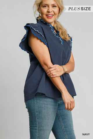 Woman wearing a navy blue sleeveless top with ruffled details and blue jeans on a gray background.
