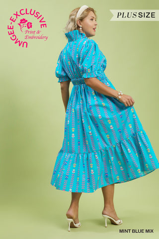 Woman wearing a blue dress with floral pattern on a green background, featuring Umgee branding.