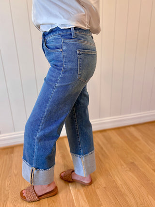 Person wearing blue jeans and a white shirt standing on a wooden floor with a white paneled wall in the background.