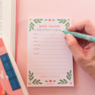 Hand writing on a 'book review' notepad with a pen on a pink background