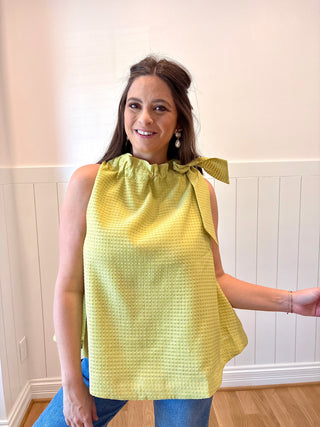 Woman wearing a yellow sleeveless top with a bow detail, standing against a white paneled wall.