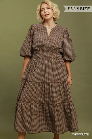 A woman modeling a brown and beige gingham tiered midi dress with puff sleeves and a split neckline, set against a solid olive green background. Text overlays read 'PLUS SIZE' at the top right and 'CHOCOLATE' at the bottom right.