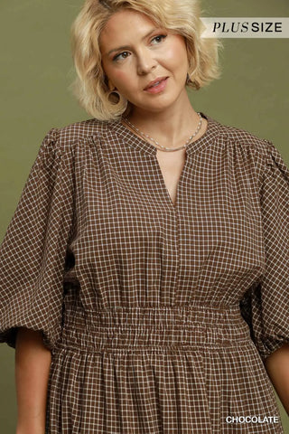 A woman wearing a brown and beige checkered plus-size dress with puff sleeves and a smocked waist, shown against a muted olive green background. Text overlays read 'PLUS SIZE' at the top right and 'CHOCOLATE' at the bottom right.