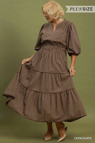 A woman modeling a brown and beige gingham tiered midi dress with puff sleeves and a smocked waist, shown against an olive green studio background. Text overlays read 'PLUS SIZE' and 'CHOCOLATE'.