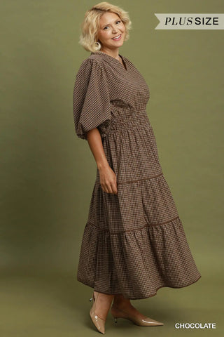 A woman modeling a brown and beige gingham tiered midi dress with puff sleeves and a smocked waist, standing against an olive green studio backdrop. Text overlays read 'PLUS SIZE' and 'CHOCOLATE'.
