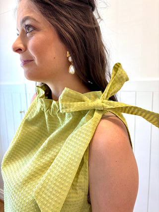 Woman wearing a green top with a bow detail, standing against a white wall.