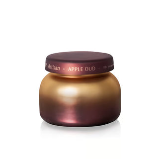 A small round cosmetic jar with a metallic gold body and a dark plum lid, featuring the text 'Artisan APPLE OUD' on the lid, set against a plain white background.