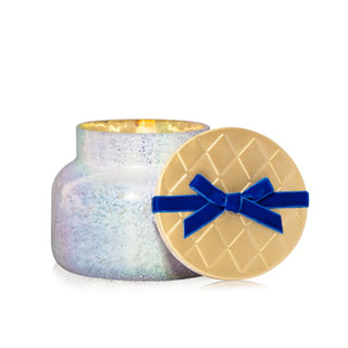 Decorative candle with iridescent glass jar and gold lid featuring a blue velvet bow on a white background.
