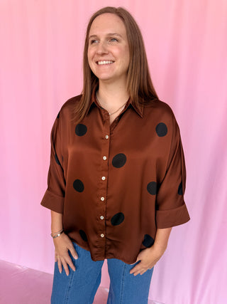 A woman with long light brown hair smiling and looking to the side, wearing a brown satin button-up blouse with large black polka dots and blue jeans, standing against a pink backdrop.