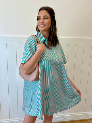 Woman wearing a blue and white striped dress holding a beige handbag against a white wall.
