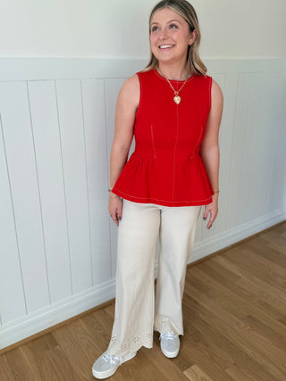 Woman wearing a red sleeveless top and white pants standing against a white paneled wall.