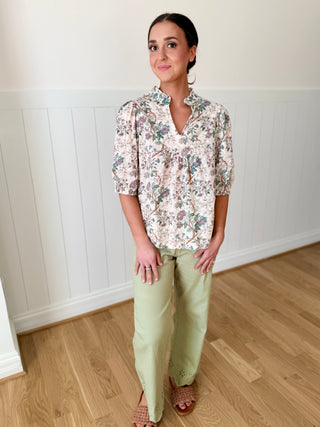 Woman wearing a floral blouse and light green pants standing in a room with wooden flooring and white walls.