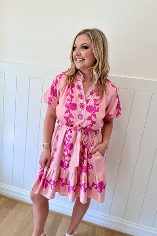 Woman wearing a pink floral dress and matching fascinator against a white paneled wall.