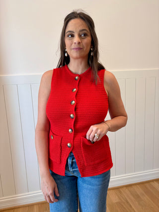 Woman wearing a red knitted vest with buttons against a white paneled wall.