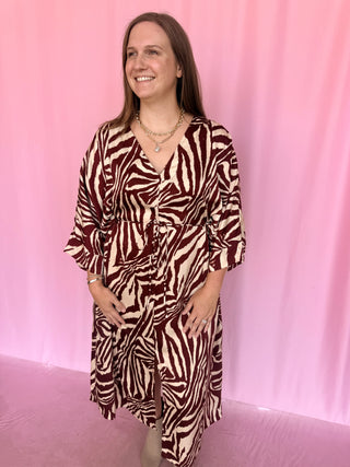 A woman wearing a brown and cream zebra-print wrap dress with three-quarter sleeves, standing against a pink backdrop.