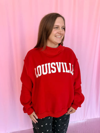 A woman wearing a bright red mock-neck sweatshirt with 'LOUISVILLE' printed in white arched letters across the chest, paired with black pants with white polka dots, standing against a pink curtain backdrop.