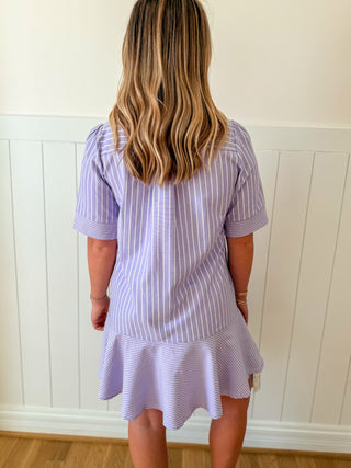 Woman wearing a purple and white striped dress against a white paneled wall.