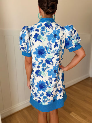 Person wearing a blue floral dress in a room with wooden floor and white walls.