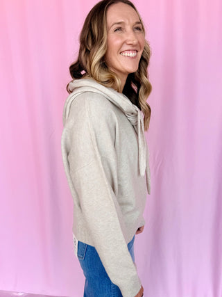 A woman with blonde-highlighted hair smiling and posing in profile, wearing a light beige knit hooded sweater and blue jeans against a pink backdrop.