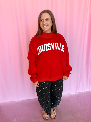 A woman wearing a red mock-neck sweatshirt with 'LOUISVILLE' printed in white arched letters across the chest, paired with black pants featuring a white polka-dot pattern and tan sandals, standing against a pink fabric backdrop.