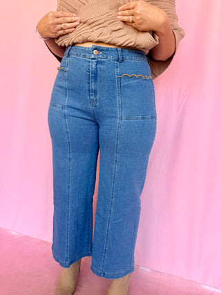 A person wearing high-waisted blue denim cropped pants with decorative zigzag stitching on the pocket flaps, paired with a beige knit top, standing against a pink backdrop.