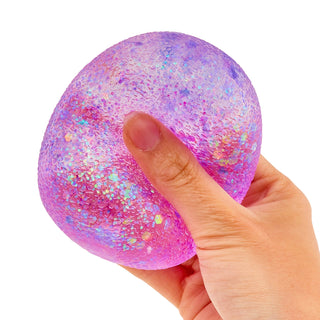A hand squeezing a round, glittery pink and purple stress ball with iridescent flecks against a plain white background.