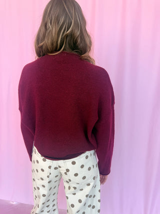A woman viewed from behind wearing a dark burgundy knit sweater and cream-colored pants with brown polka dots, standing against a pink draped backdrop.