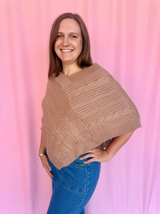 A woman wearing a chunky knit poncho sweater in a warm taupe-brown color, paired with blue jeans, standing against a pink fabric backdrop.