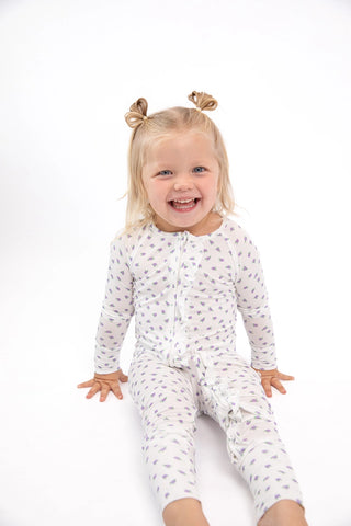 A smiling young girl with blonde hair in two buns sits on a white studio background wearing a white long-sleeve footless pajama romper with a small gray leaf pattern.