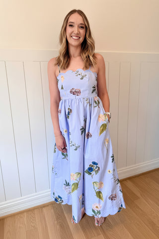 Woman wearing a floral dress with a blue fascinator against a white paneled wall.