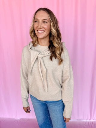 A woman with blonde-highlighted hair smiling, wearing a light beige knit sweater with a knotted tie detail at the neck and blue jeans, standing against a pink fabric backdrop.