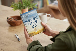 Person holding a book titled 'Never Give Up' by Max Lucado on a wooden table.