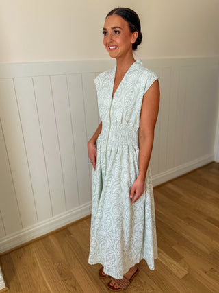 Woman wearing a light green lace dress standing in a room with wooden flooring and white walls.