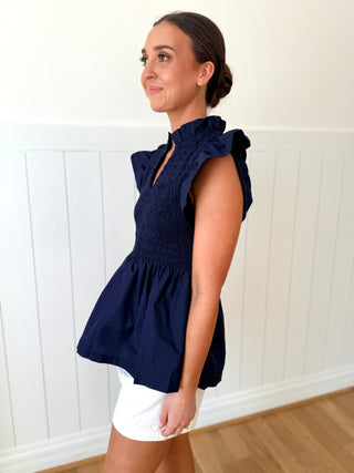 Woman wearing a navy blue top with ruffled sleeves and white shorts against a white paneled wall.