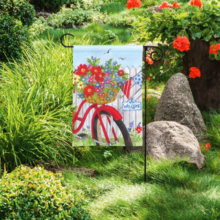 Garden flag with bicycle and flowers in a basket on a garden background with rocks and flowers.