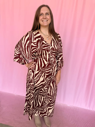 A woman modeling a long-sleeved, wrap-style maxi dress with a bold zebra print in dark red and cream, standing against a pink backdrop.
