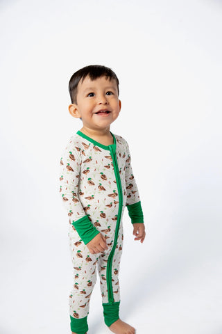 A toddler standing against a plain white studio background, wearing a long-sleeve footless pajama top with green trim and a small holiday-themed print.