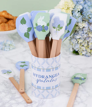 Set of spatulas with floral designs in a canister labeled 'Hydrangea Spatulas' on a light surface with flowers in the background.