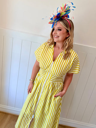 Woman wearing a yellow and white striped dress with a colorful headpiece against a white paneled wall.