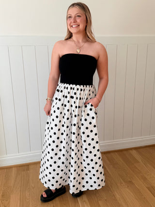 Woman wearing a black strapless top and white polka dot pants standing in a room with wooden flooring and white paneled walls.