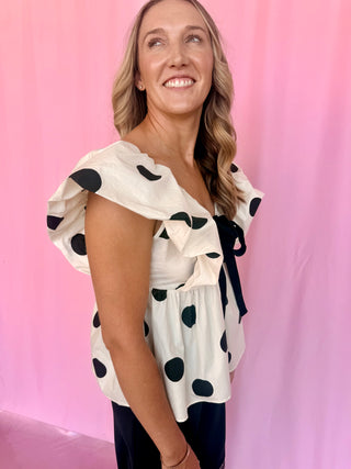 A woman wearing a white ruffled blouse with black polka dots and a black bow tie, standing against a pink backdrop.