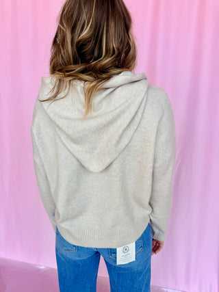 A woman viewed from behind wearing a light beige knit hooded sweater and blue jeans, standing against a pink backdrop.