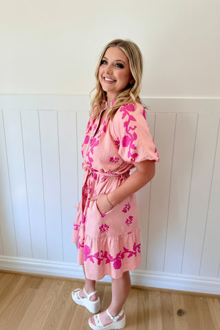 Woman wearing a pink floral dress and matching headpiece against a white paneled wall.