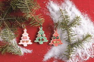 Three Christmas tree-shaped ornaments in pink, mint green, and coral red with holiday details, arranged on a red wooden surface with pine branches and white faux snow.
