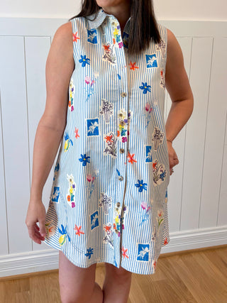 Woman wearing a sleeveless dress with colorful floral patterns against a white paneled wall.