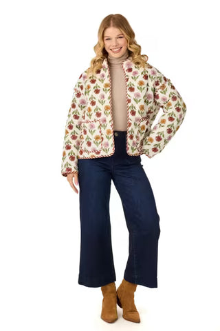 A woman modeling a cream-colored quilted jacket with a pink and green floral pattern and striped trim, paired with a beige turtleneck, dark blue wide-leg jeans, and tan ankle boots, against a solid white background.