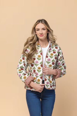 A woman modeling a white bomber jacket with a colorful floral pattern and red-and-white striped ribbed cuffs and hem, paired with blue jeans against a solid beige background.