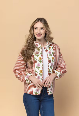A woman modeling a long-sleeve button-up shirt with red and white vertical stripes on the sides and a white front panel decorated with a colorful floral print, paired with a white top and blue jeans, against a solid beige background.
