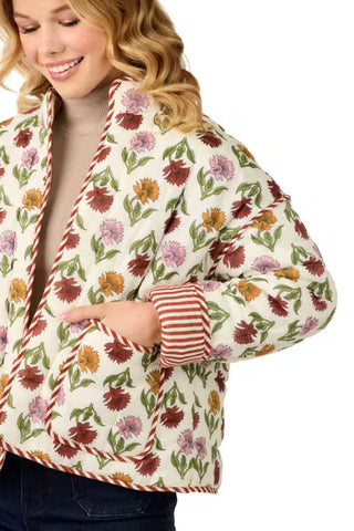 A woman wearing a cream-colored quilted jacket with a colorful floral print and striped trim, paired with a beige turtleneck and dark jeans, shown against a plain white background.